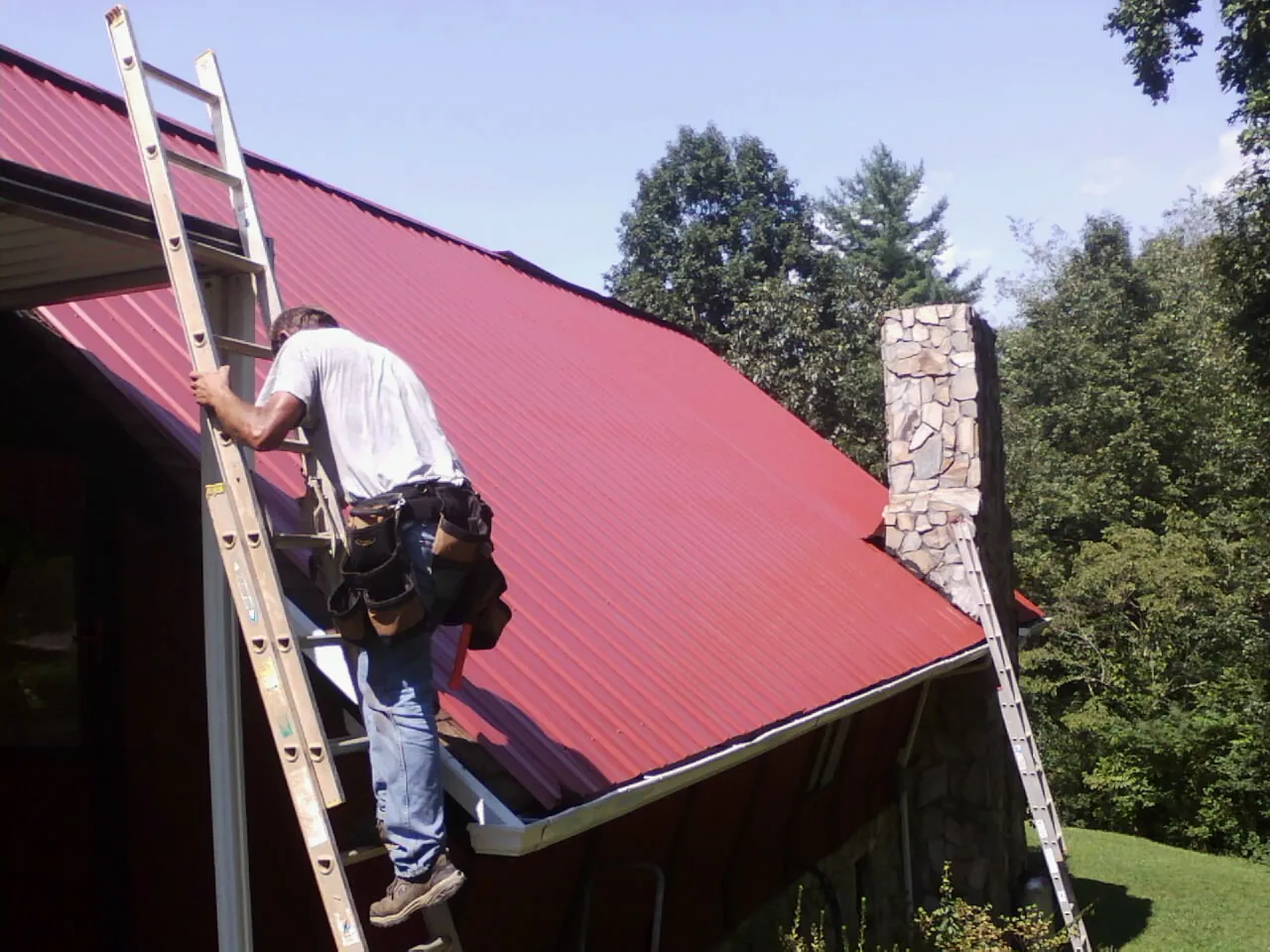 Roofer on a ladder beside a red metal roof and stone chimney — Soffit & Fascia Repair by Roofing Brothers in North Bay